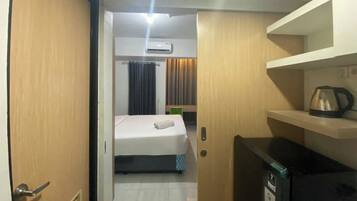 A hotel room with a bed, a small kitchenette, a shelf with a kettle, and an air conditioning unit.