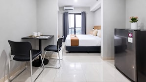 Quarto de hotel com cama, escrivaninha com cadeiras, geladeira e janela com cortinas.