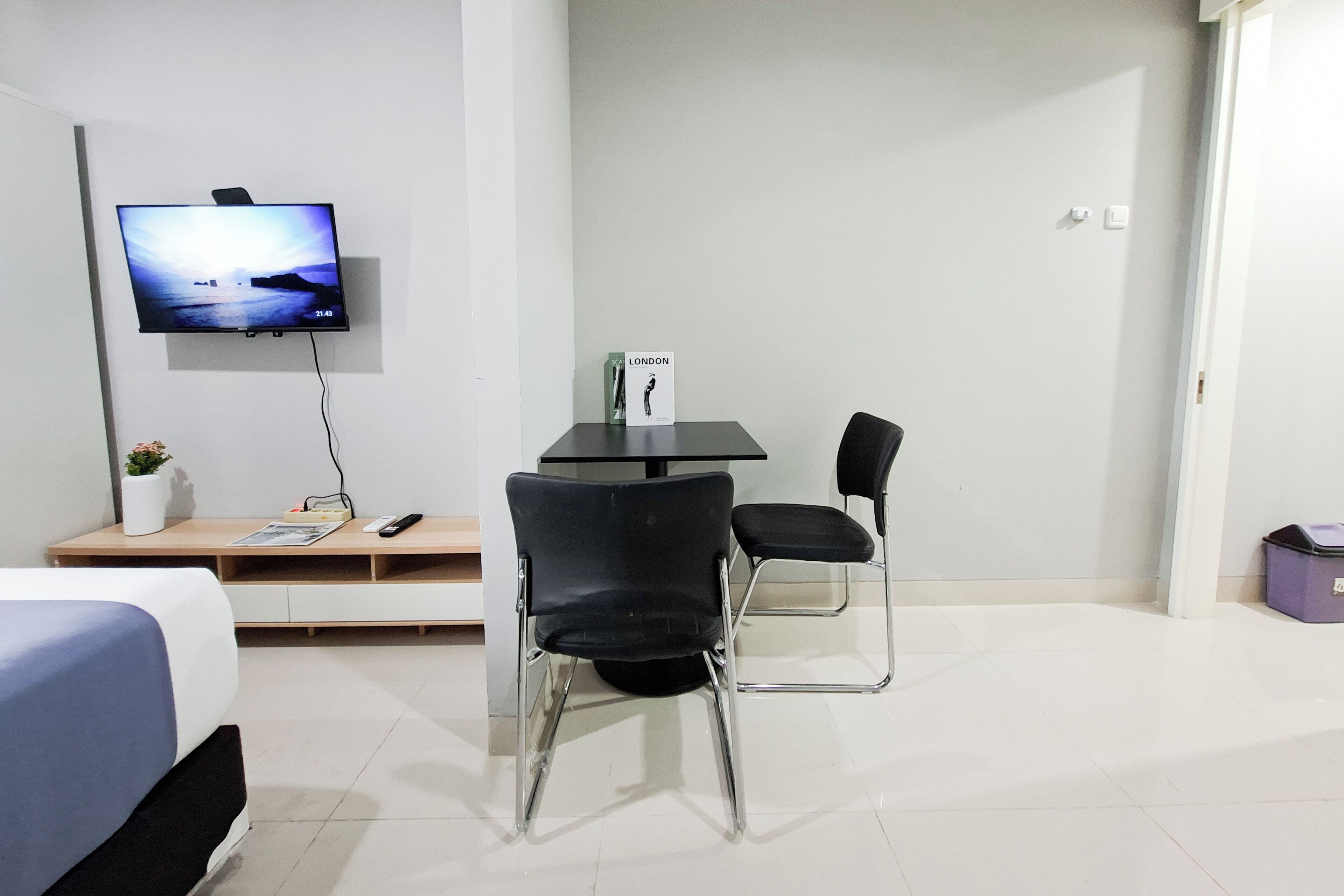 A hotel room with a bed, a TV mounted on the wall, a small table with chairs, and a bedside table with a remote control and a plant.
