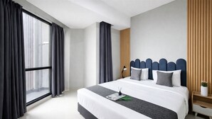 Una habitación de hotel moderna con una cama grande, un cabecero de madera, una mesita de noche con lámpara y una ventana con cortinas.