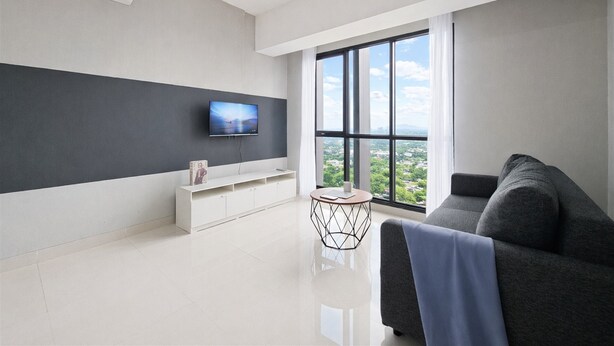 A modern living room with a flat-screen TV, a sofa, a coffee table, and a large window with a city view. - Homey And Spacious 3Br With City View At Apartment The Ayoma (South Tangerang)