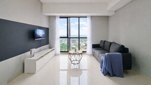 A modern living room with a sofa, a TV mounted on the wall, a small round table, and a large window offering a city view. - Homey And Spacious 3Br With City View At Apartment The Ayoma (South Tangerang)