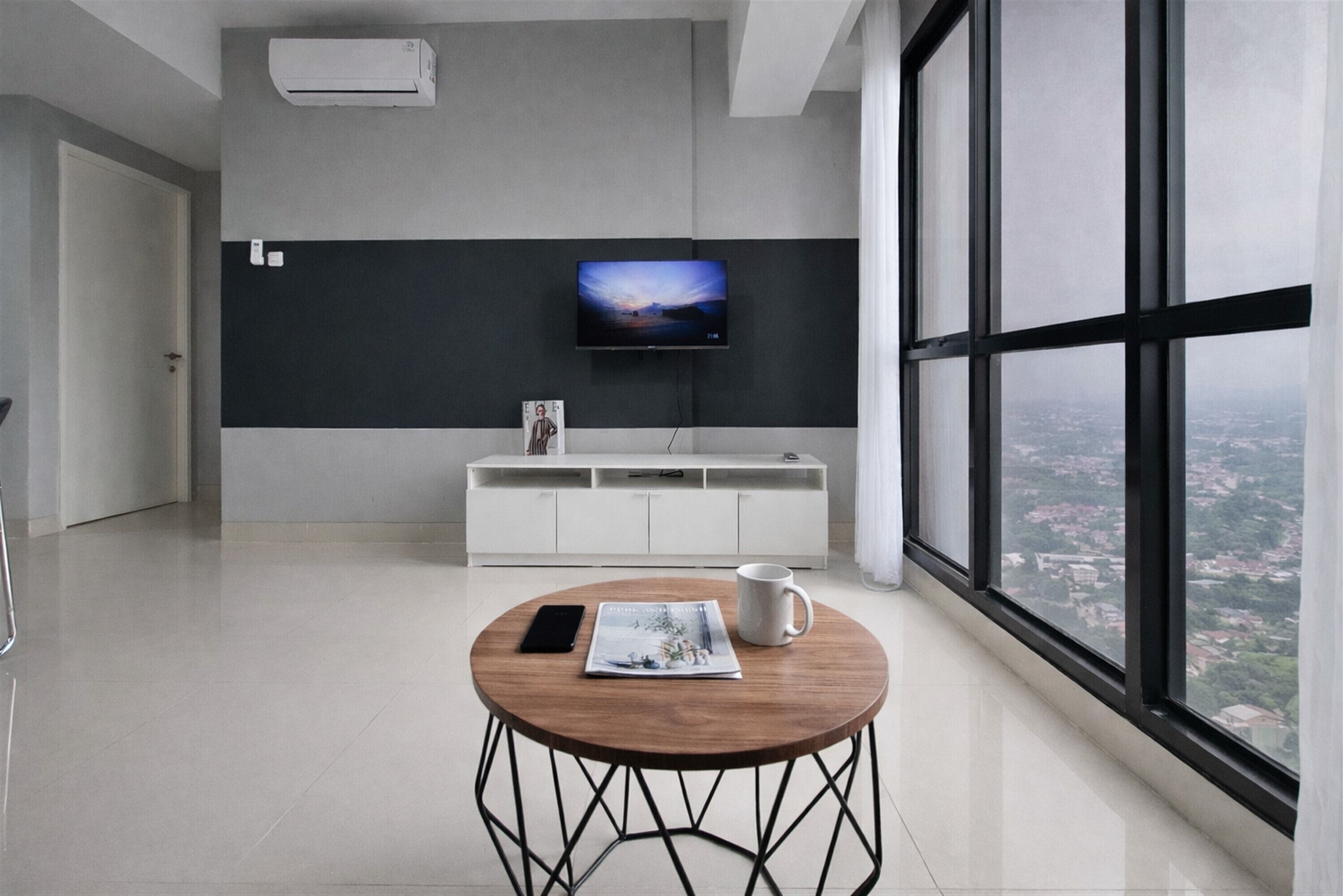 A modern room with a large window, a flat-screen TV mounted on the wall, a white cabinet, and a round table with a magazine and a mug.