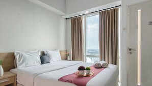 A hotel room with a bed, a nightstand, a window with curtains, and a view of the city. - Cozy Living Studio At The Square Surabaya Apartment (Surabaya)