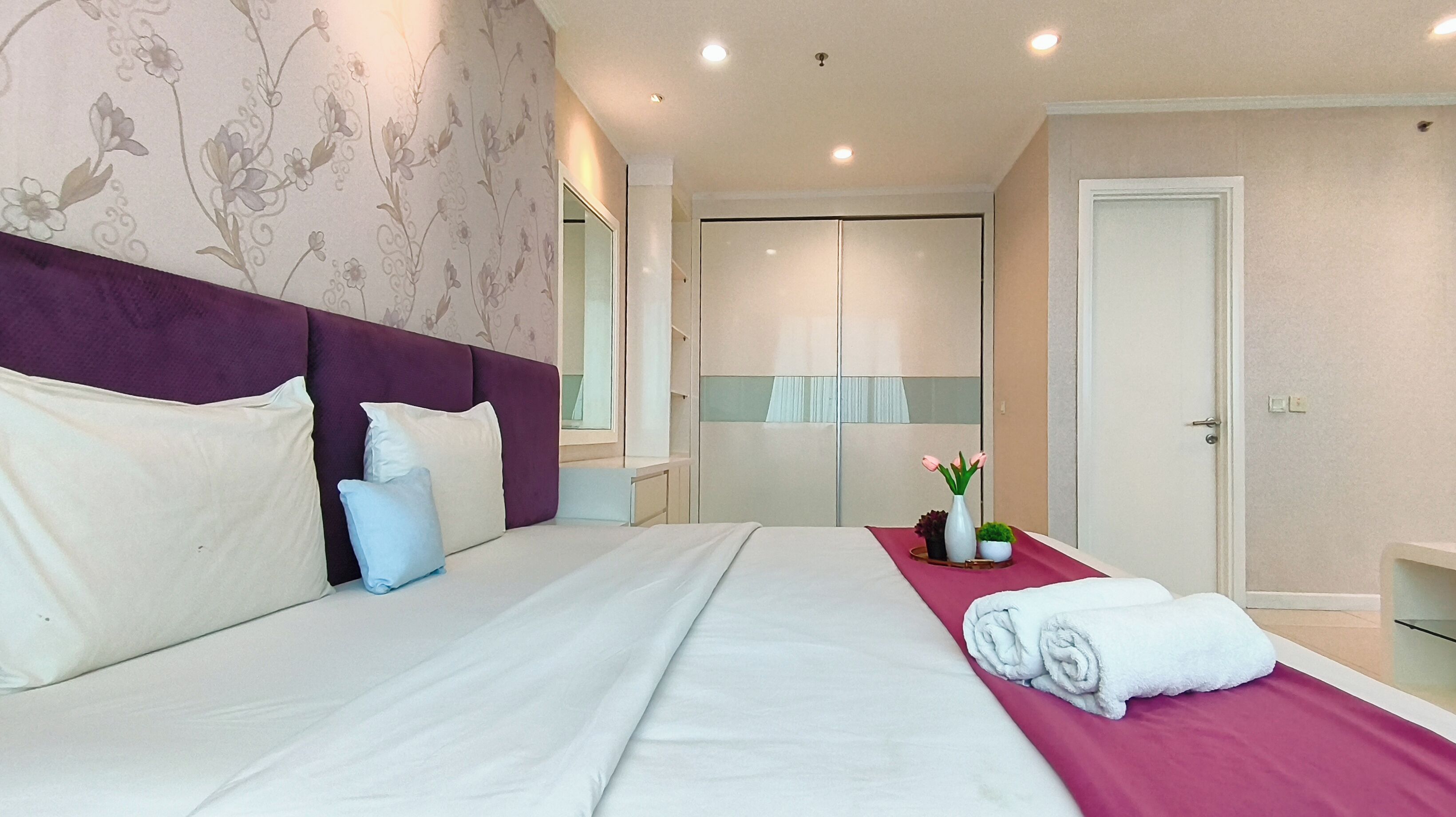 Moderni hotellihuone, jossa on suuri sänky, violetti pääty, peili, meikkipöytä ja ovi.