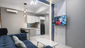 Una sala de estar moderna con un sofá, un televisor montado en la pared y una cocina al fondo.