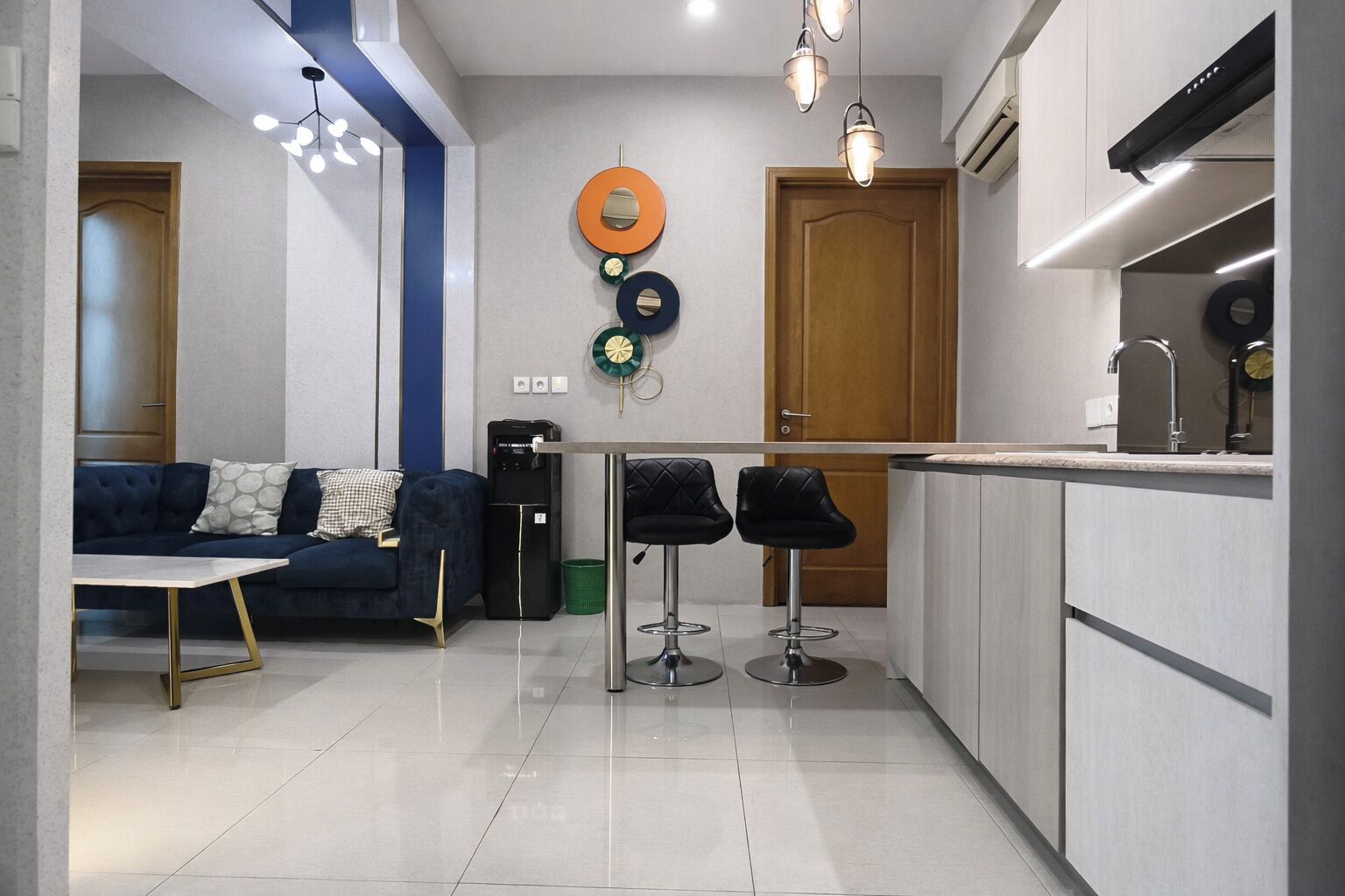A modern kitchen with a built-in oven, a dining area with a sofa and two bar stools, and a wall-mounted light fixture.