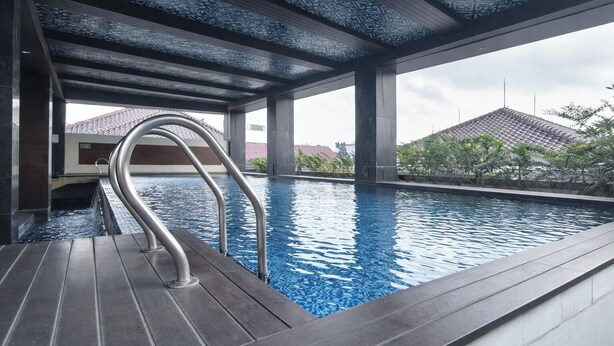 Una piscina interior con una terraza de madera, escalera metálica y vistas a edificios con tejados de tejas.