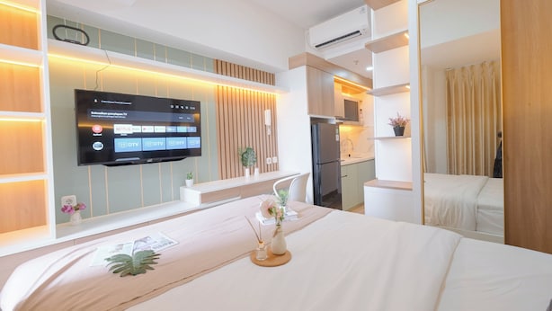 A hotel room with a bed, a TV mounted on the wall, a small table with a vase, and a kitchen area. - Warm Minimalist Studio Room At Tokyo Riverside Pik 2 Apartment (Teluknaga)