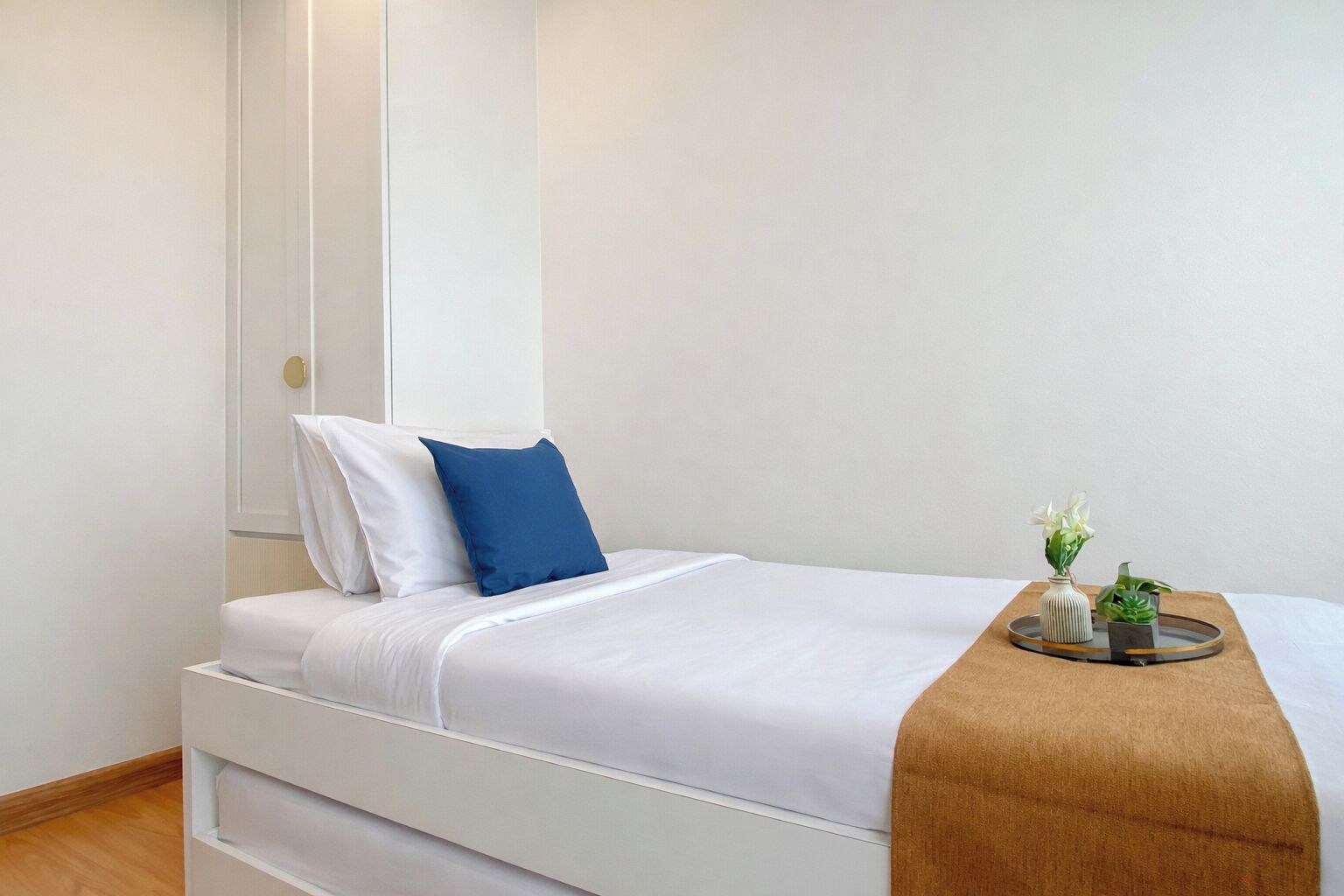 A single bed with white bedding, a blue pillow, and a brown throw blanket. There is a small table with a vase and plants.