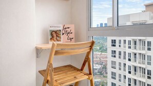 Rincón de una habitación con una silla de madera, una pared blanca, una ventana con vista a la ciudad y un estante con un libro y un cartel.