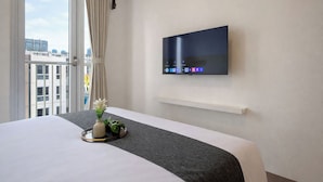 Una habitación de hotel con cama, televisor montado, ventana con cortinas y vista a edificios.