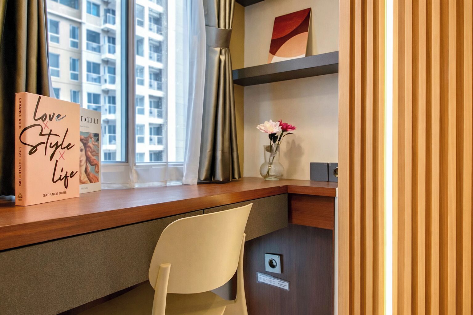 Una habitación de hotel con un escritorio, una silla, un libro titulado 'Love Style Life', un jarrón con flores y vistas a edificios a través de la ventana.