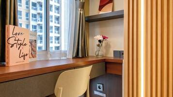 Una habitación de hotel con un escritorio, una silla, un libro titulado 'Love Style Life', un jarrón con flores y vistas a edificios a través de la ventana.
