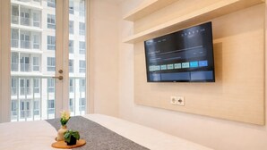 Ein Hotelzimmer mit einem an der Wand befestigten Fernseher, der den Film 'The Family Plan 2' zeigt, einem Bett mit grauem Teppich, einer kleinen Pflanze auf dem Bett und einem Fenster mit Blick auf ein Gebäude.
