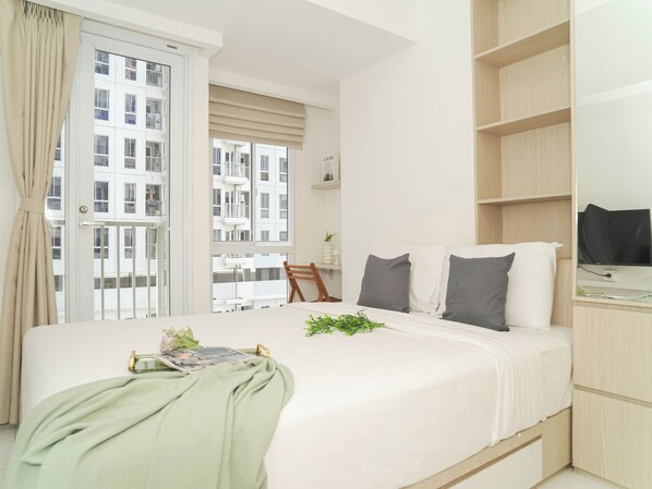 A modern hotel room with a large bed, a TV, a bookshelf, and a view of the cityscape. - Strategic And Stylish Studio At Apartment Tokyo Riverside Pik 2 (Teluknaga)