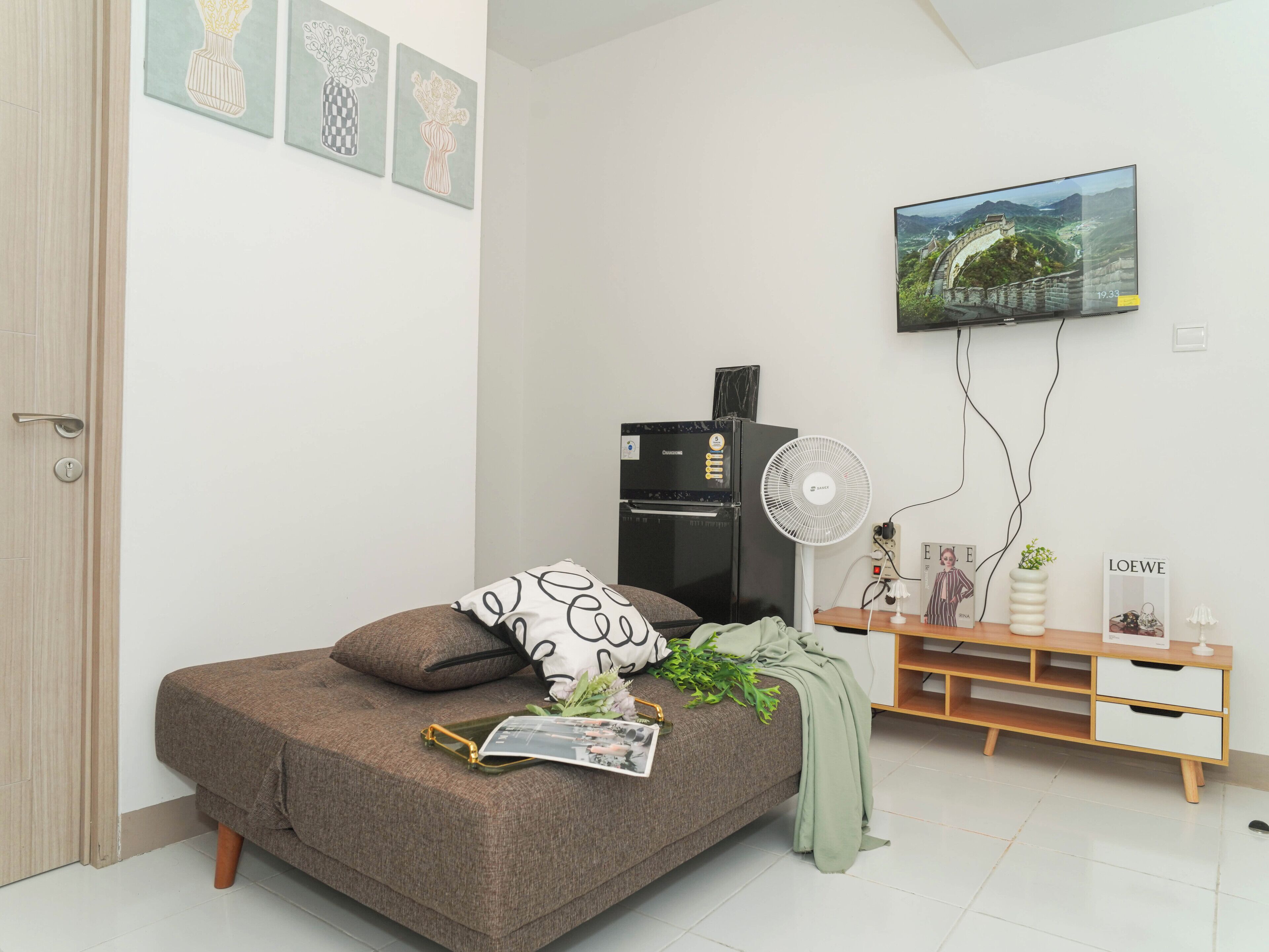 A bedroom with a bed, a wall-mounted TV, a refrigerator, a fan, and a wooden side table with decorative items.