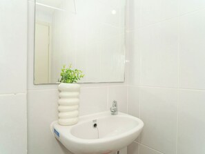 A white bathroom sink with a mirror above it, a white vase with green plants, and white tiled walls. - Functional 2Br For 3 Pax At Tokyo Riverside Pik 2 Apartment (Teluknaga)
