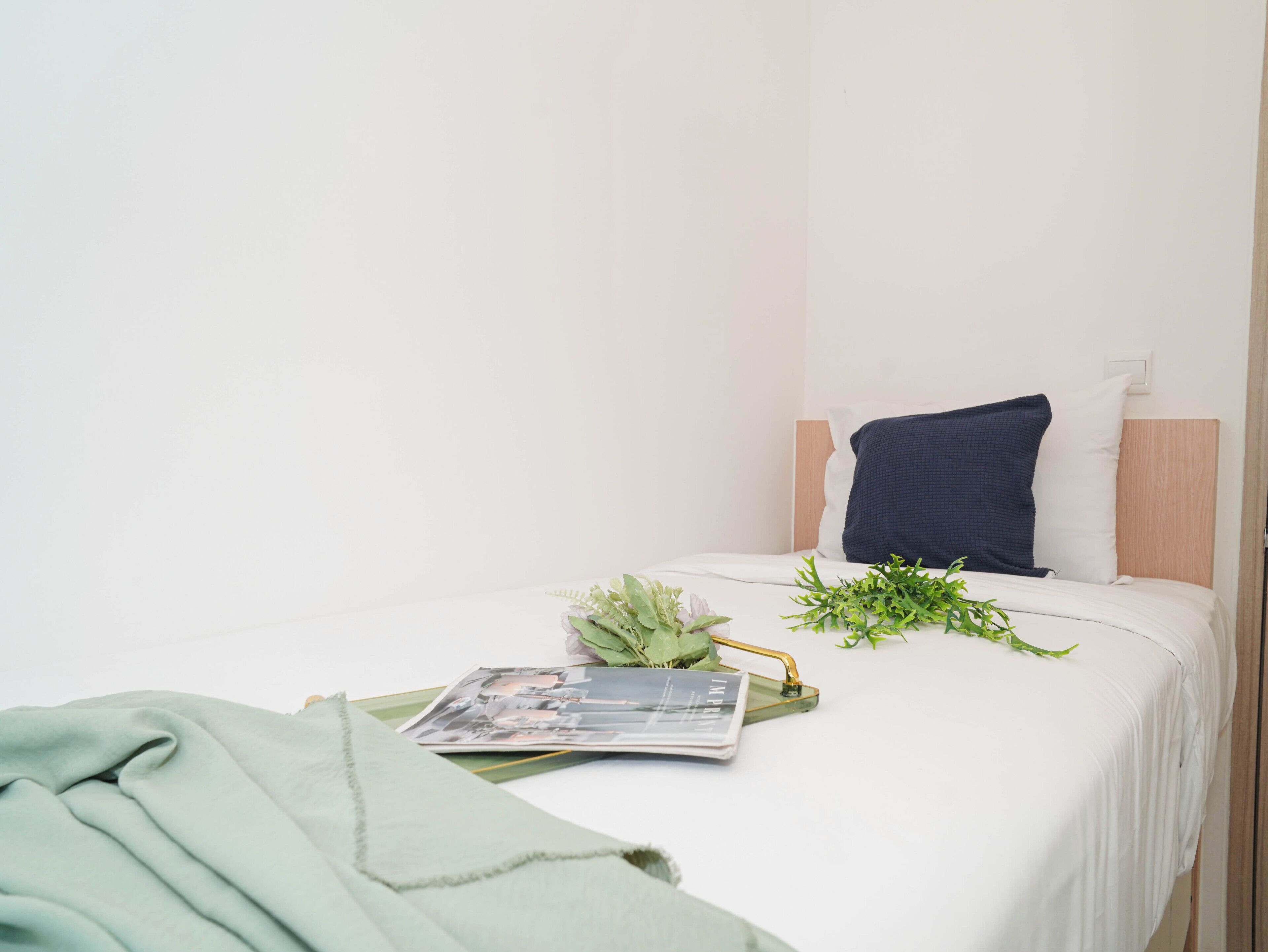 A neatly made bed with a magazine, a green cloth, and a plant on the side.