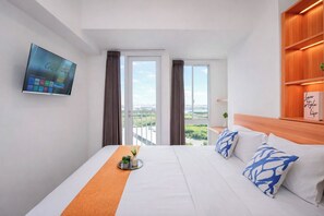 A hotel room with a bed, a TV, a view of the city, and a shelf with a book. - Neat And Minimalist Studio At Tokyo Riverside Pik 2 Apartment Near Tokyo Hub (Teluknaga)