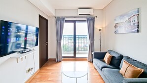 A living room with a flat-screen TV, a glass coffee table, a sofa with cushions, a painting on the wall, and a view through a large window. - Cozy Living 3Br At Transpark Bintaro Apartment (South Tangerang)
