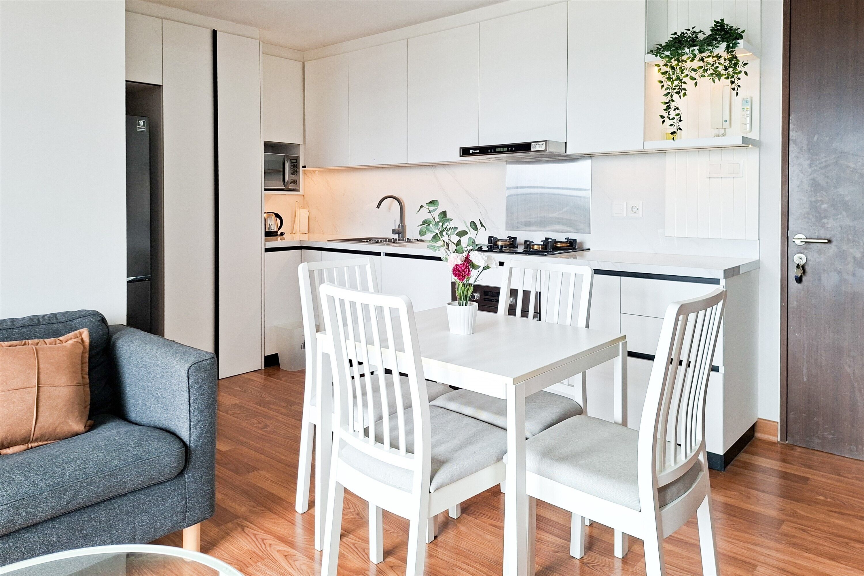 A modern kitchen with white cabinetry, a dining area with a white table and chairs, and a grey sofa with a brown cushion.