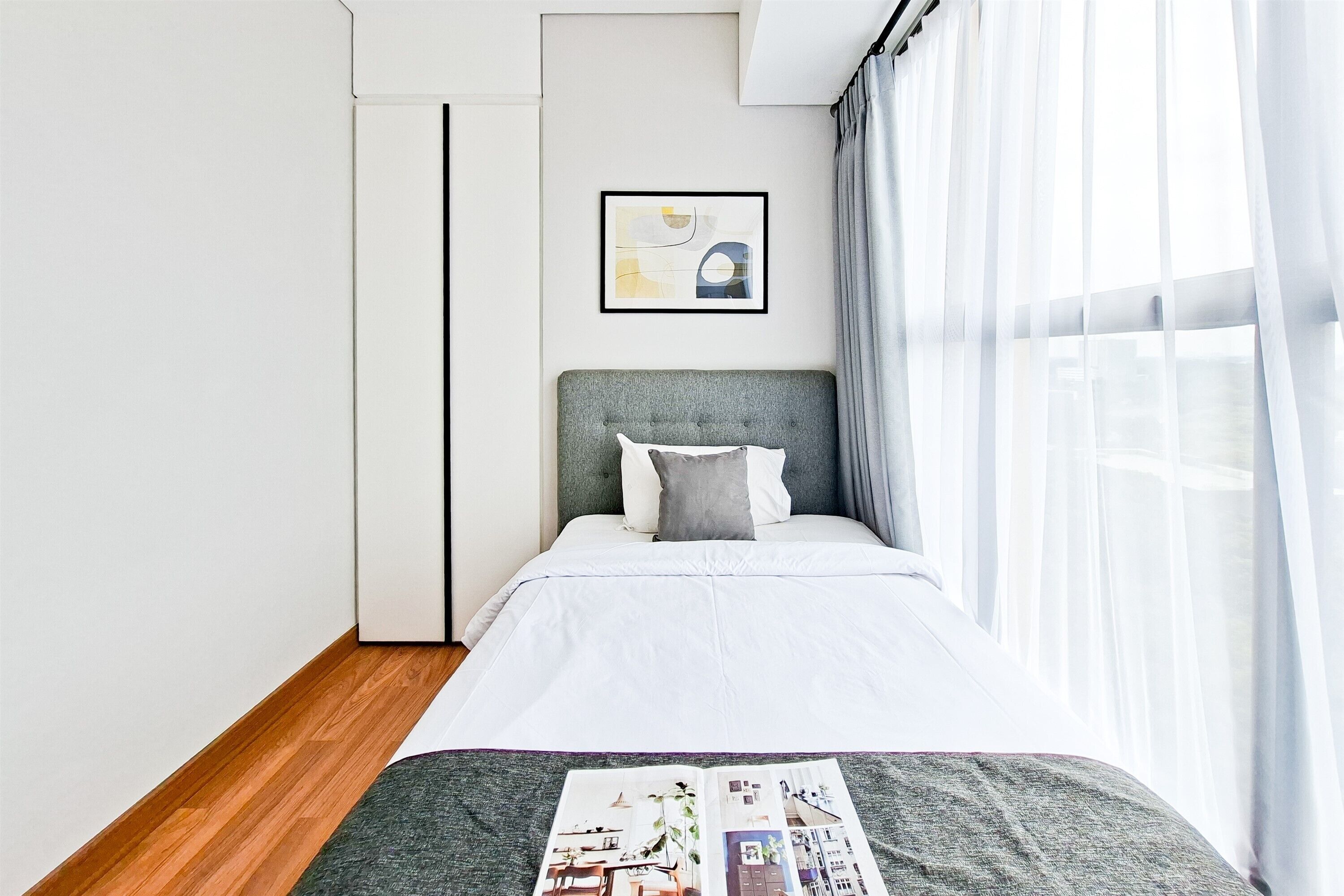 Un dormitorio moderno con cabecera gris, ropa de cama blanca, una obra de arte abstracta enmarcada y una ventana con cortinas translúcidas.