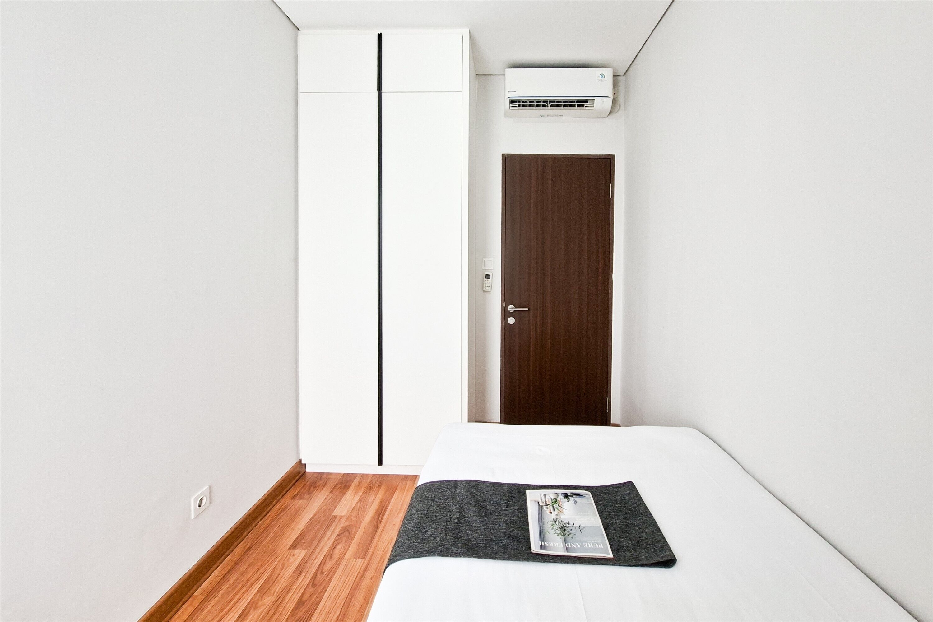 A bedroom with a bed, a wooden floor, a white wardrobe, a dark door, and an air conditioning unit.