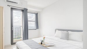 Moderni hotellihuone, jossa on suuri sänky, ikkuna verhoilla, näkymä kaupunkiin sekä pieni pöytä, jossa on maljakko ja kirja.