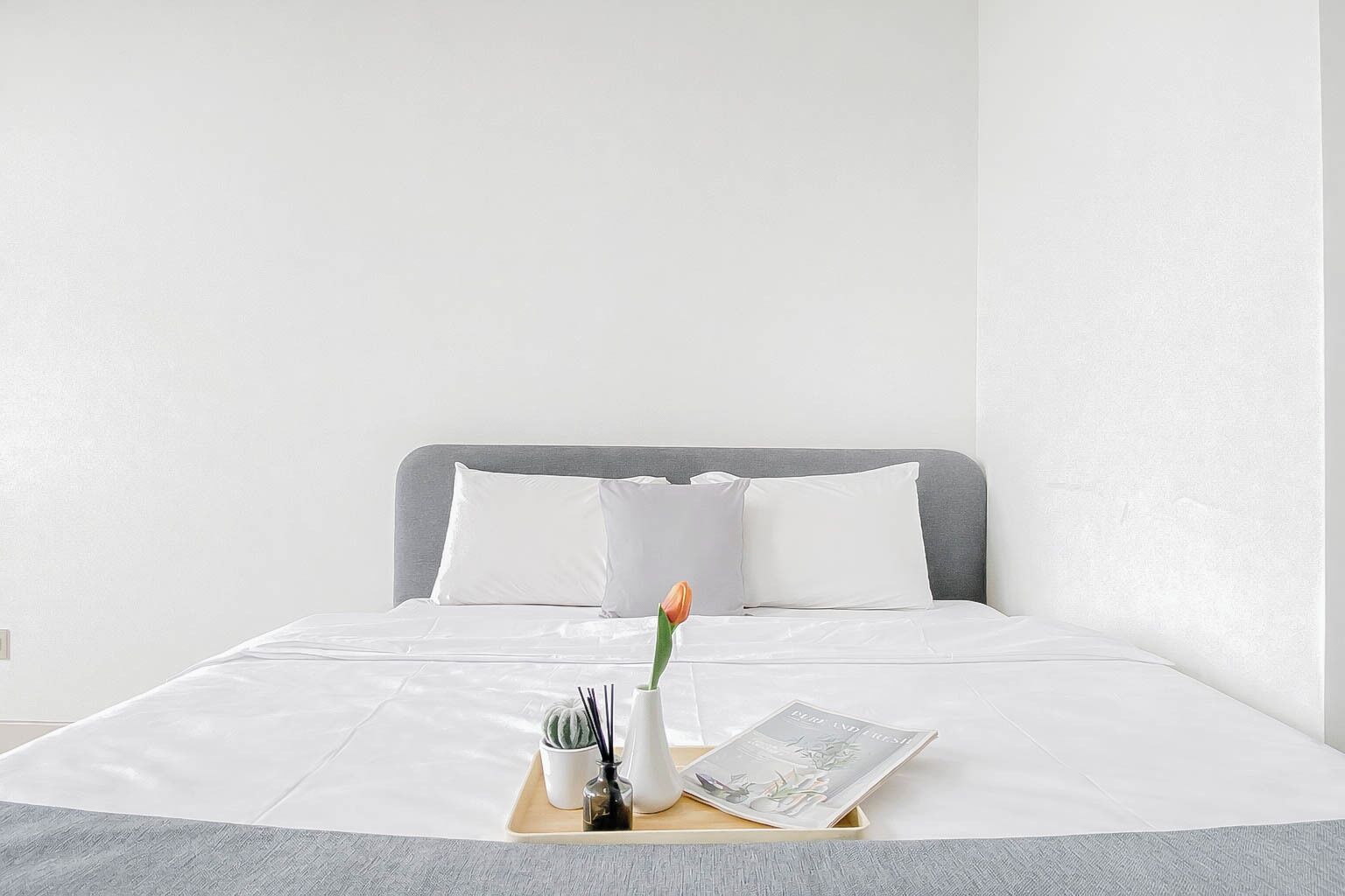 A neatly made bed with white linens, a gray headboard, and a tray with a flower, a small cactus, and a magazine.