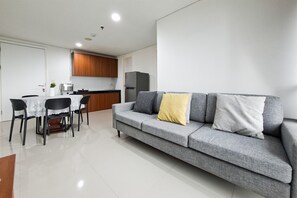 A modern living area with a grey sofa, a dining table with chairs, and a kitchen in the background. - Comfy White 2Br For 3 Pax At Tuscany Residence (South Tangerang)