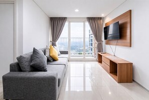 A modern living room with a grey sofa, wooden TV stand, and a balcony with curtains. - Comfy White 2Br For 3 Pax At Tuscany Residence (South Tangerang)