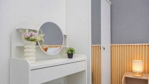 Un dormitorio con un tocador blanco, un espejo redondo, una pequeña planta en maceta, una mesita de noche con lámpara y un revestimiento de madera en listones.