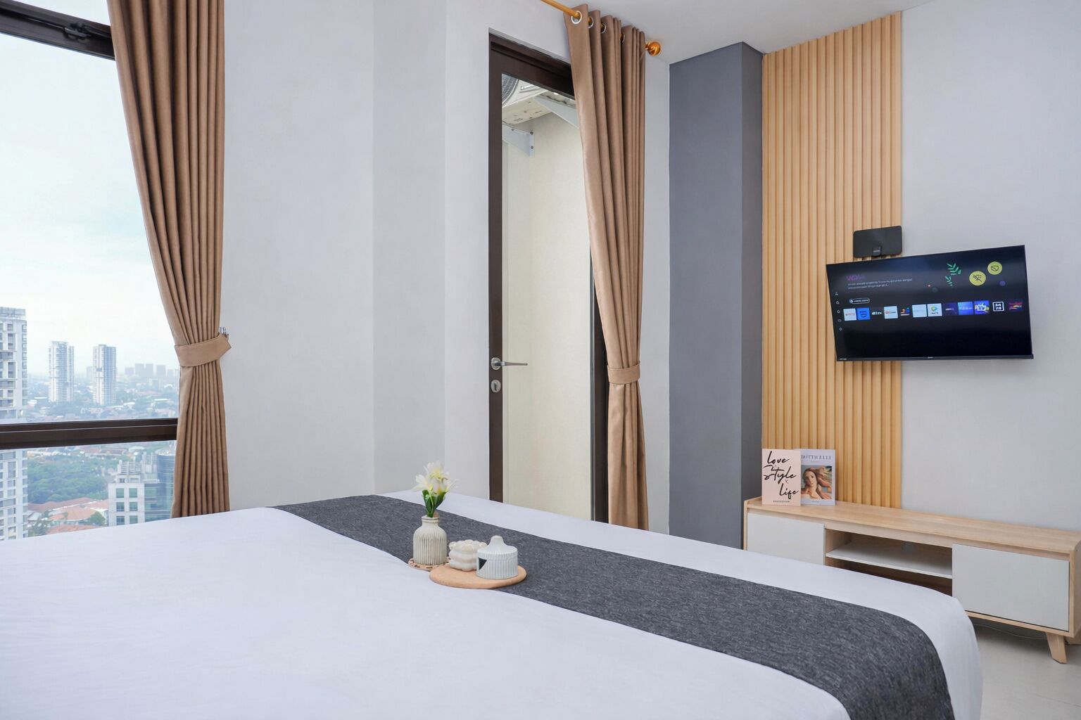 A hotel room with a bed, a TV on the wall, a window with curtains, and a view of the cityscape.