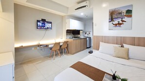 A hotel room with a bed, a desk with chairs, a TV, a kitchen area, and a painting on the wall. - Wooden Touch Restful Studio At Apartment West Vista (Jakarta)