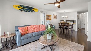 A living room with a sofa, a coffee table, a ceiling fan, and a dining area with chairs and a table. - Harborside 201: Charming Condo Steps from the Beach (Myrtle Beach)