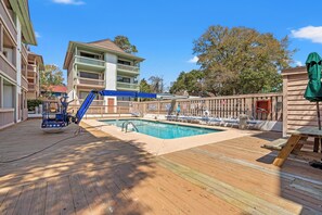 A pool area with a wooden deck, lounge chairs, and a building with balconies. - Harborside 201: Charming Condo Steps from the Beach (Myrtle Beach)