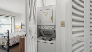 A compact room with a bed, a washing machine, and a small window. - Harborside 201: Charming Condo Steps from the Beach (Myrtle Beach)