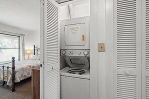 A compact room with a bed, a washing machine, and a small window. - Harborside 201: Charming Condo Steps from the Beach (Myrtle Beach)