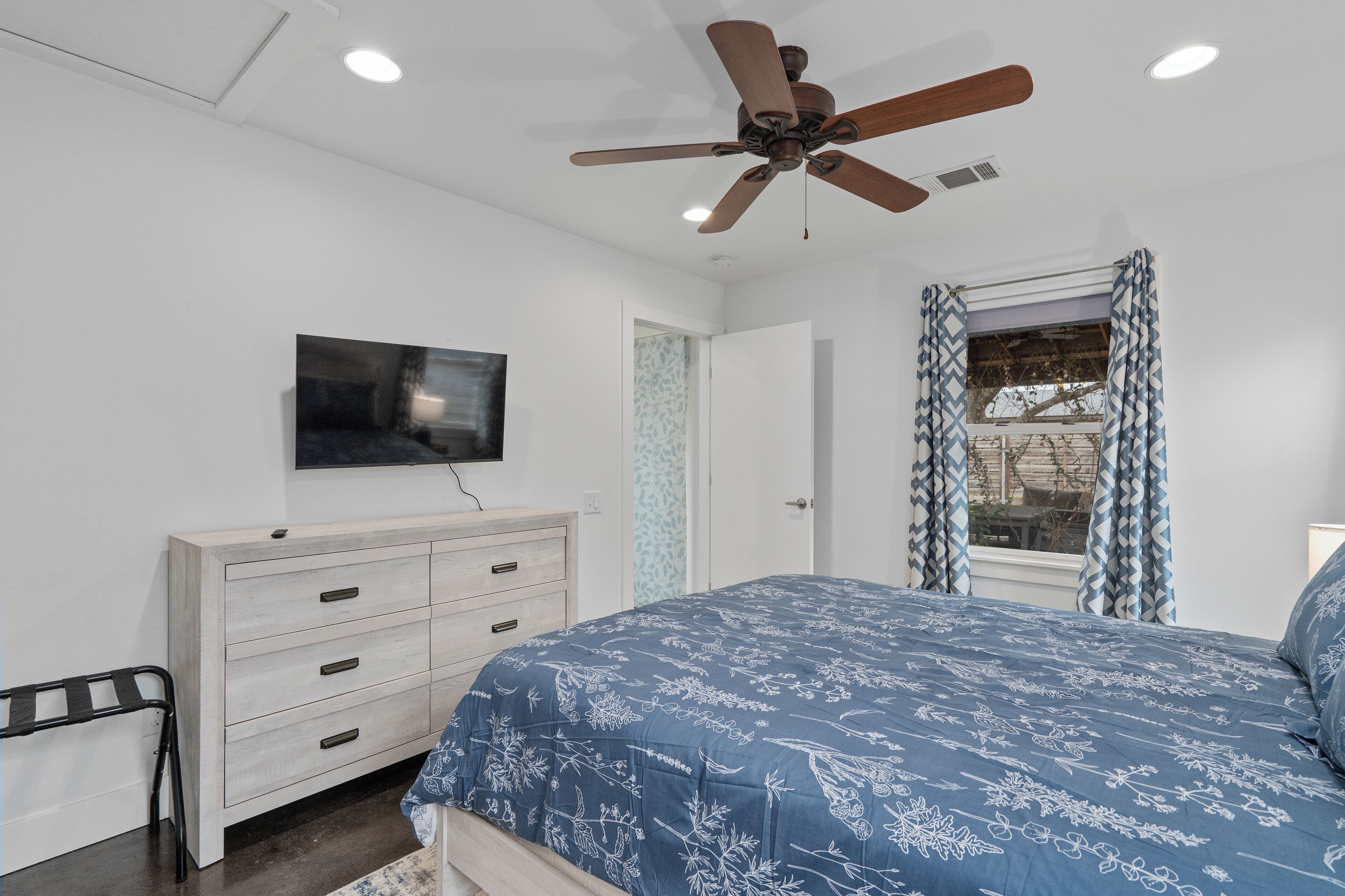 A bedroom with a bed, a ceiling fan, a TV mounted on the wall, a dresser, and a window with patterned curtains.