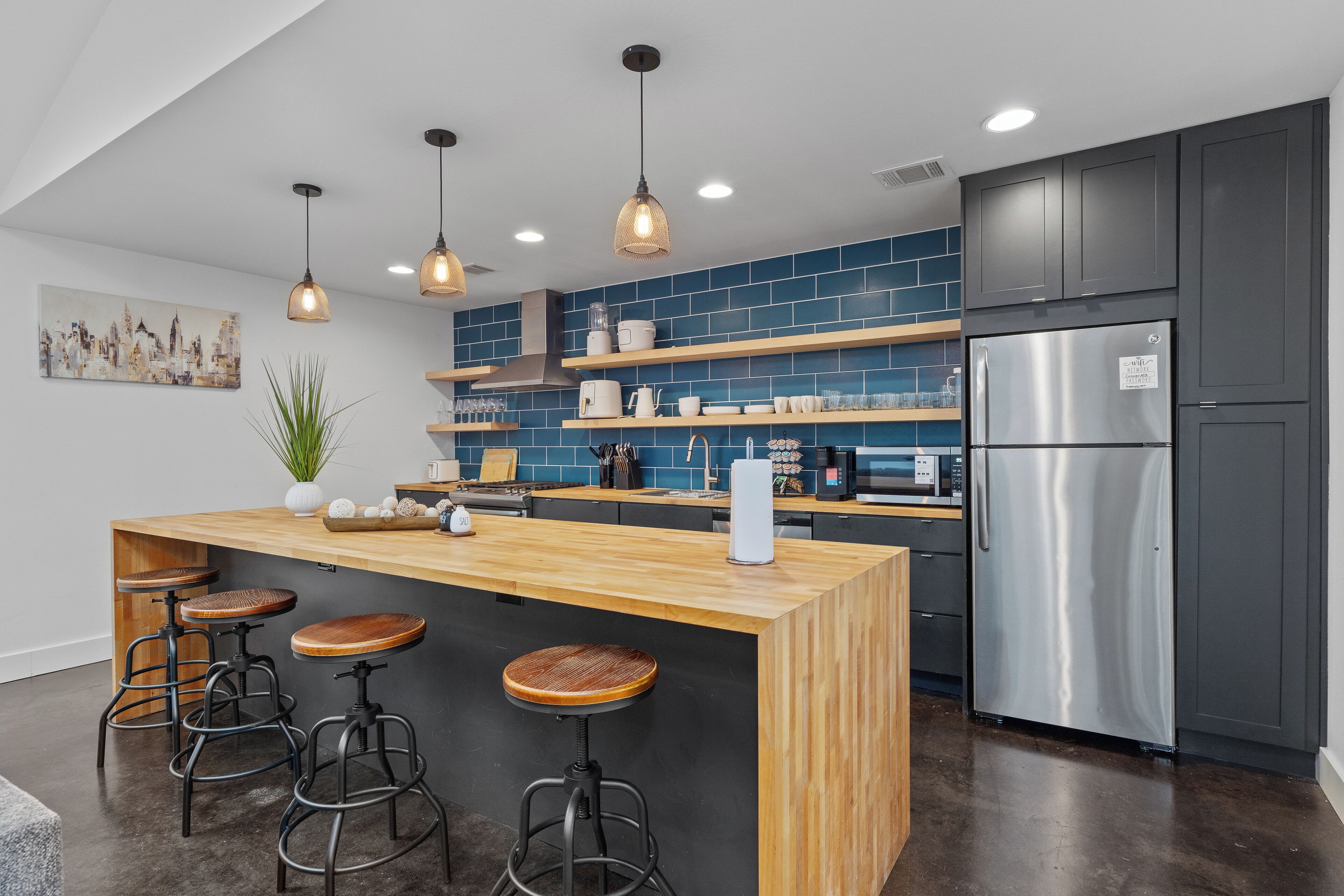A modern kitchen with a central island, bar stools, a stainless steel refrigerator, and blue tiled backsplash.