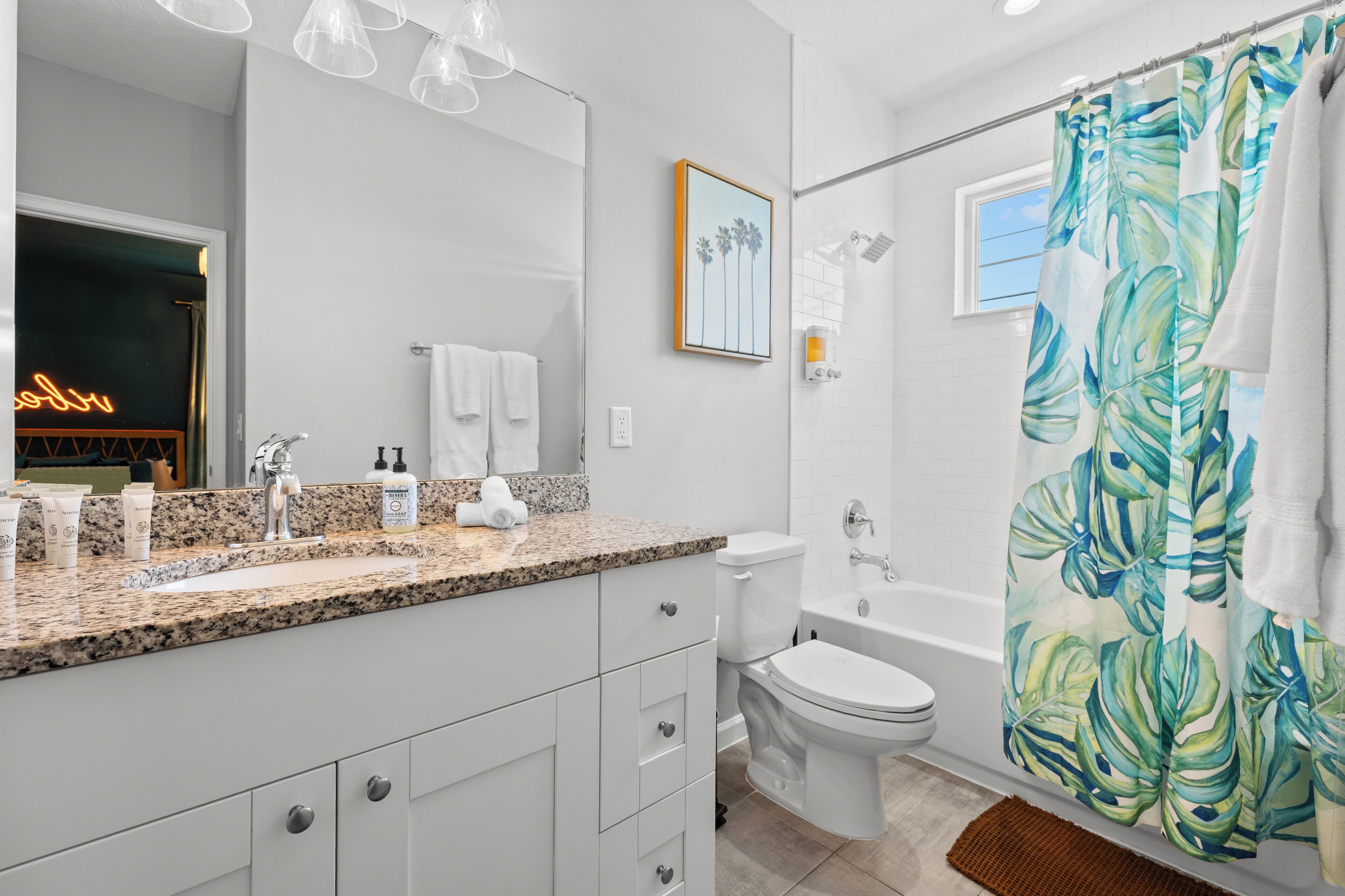Un baño moderno con encimera de granito, accesorios blancos, cortina de ducha con motivos de hojas tropicales y un cuadro en la pared.