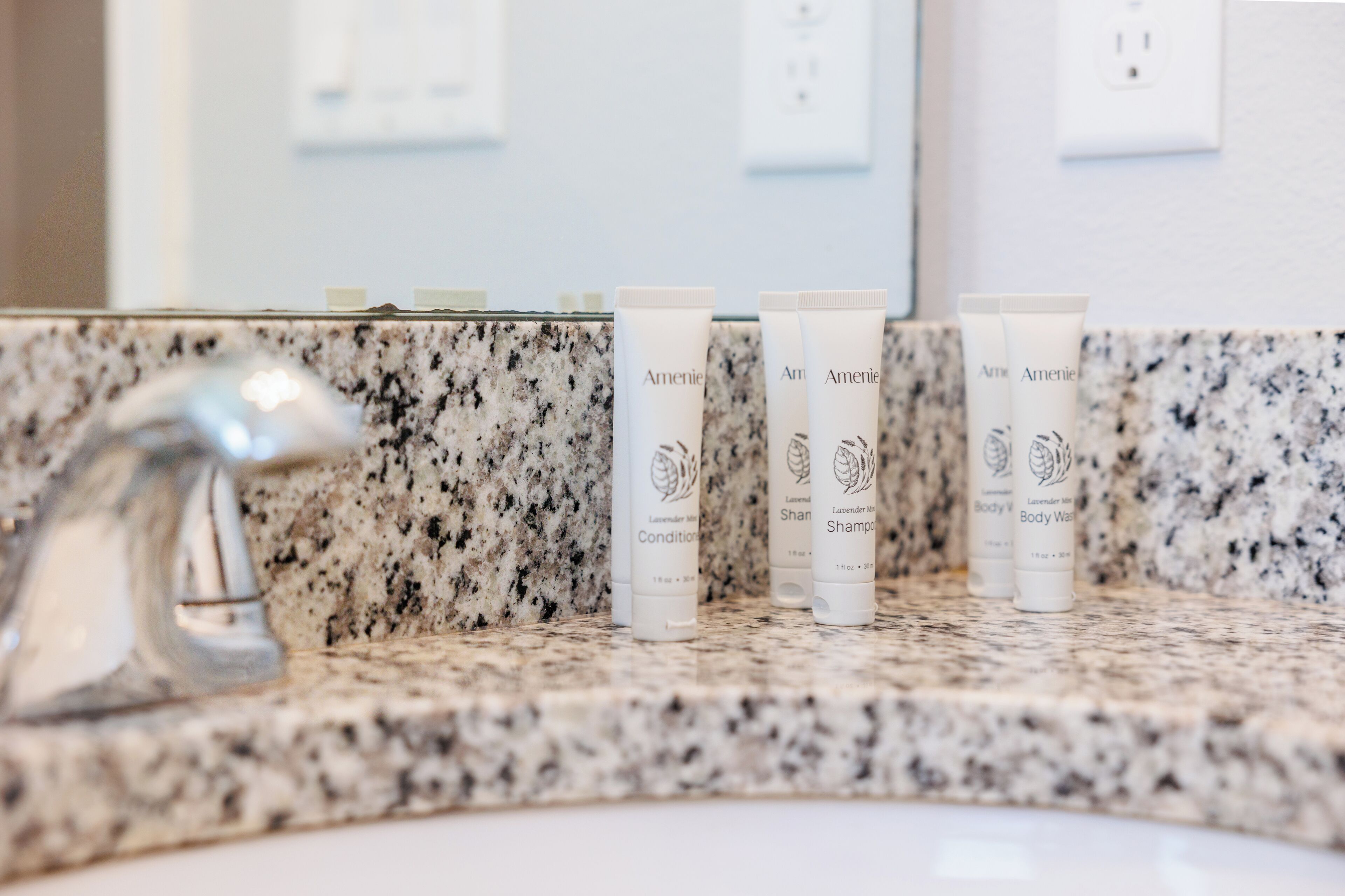 A bathroom with a granite countertop and a silver faucet. There are four tubes of body wash and shampoo on the counter.