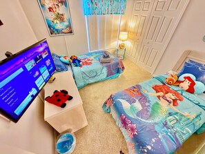 Ein Kinderzimmer mit Fernseher, zwei Betten mit Disney-Motiven, einem kleinen Tisch und einem Fenster mit Vorhängen.