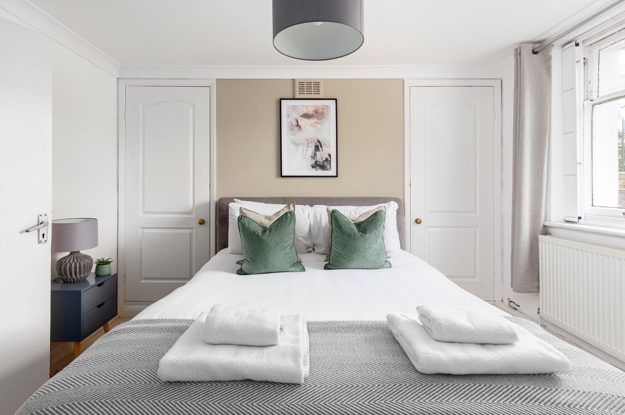 A neatly arranged bedroom with a bed, pillows, a bedside table, a lamp, and a framed artwork on the wall.