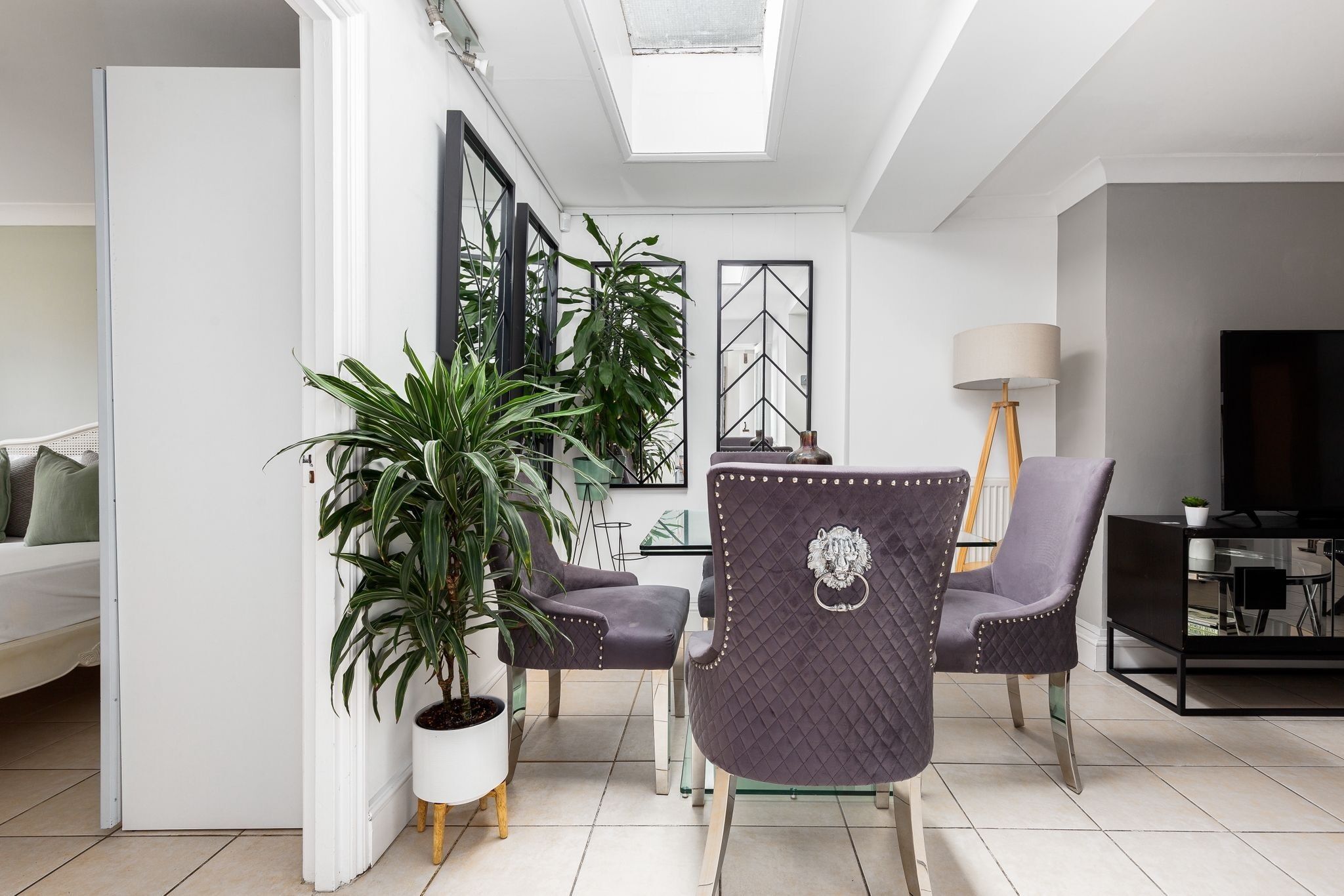 A modern living room with a dining area, featuring a large potted plant, two upholstered chairs, a glass dining table, and a television.