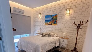 Een slaapkamer met een witte baksteenmuur, een bed met witte beddengoed, een schilderij, een nachtkastje met een lamp en een kapstok.
