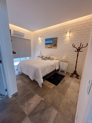 A bedroom with a white brick wall, a bed with white bedding, a painting, a nightstand with a lamp, and a coat rack. - Juquehy Corner (São Sebastião)