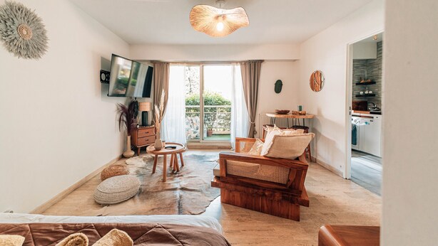 Ein großzügiger Wohnbereich mit einer Couch, einem kleinen runden Tisch, einem Sessel und einem Esstisch. Ein großes Fenster lässt Tageslicht herein und bietet einen Blick ins Freie.