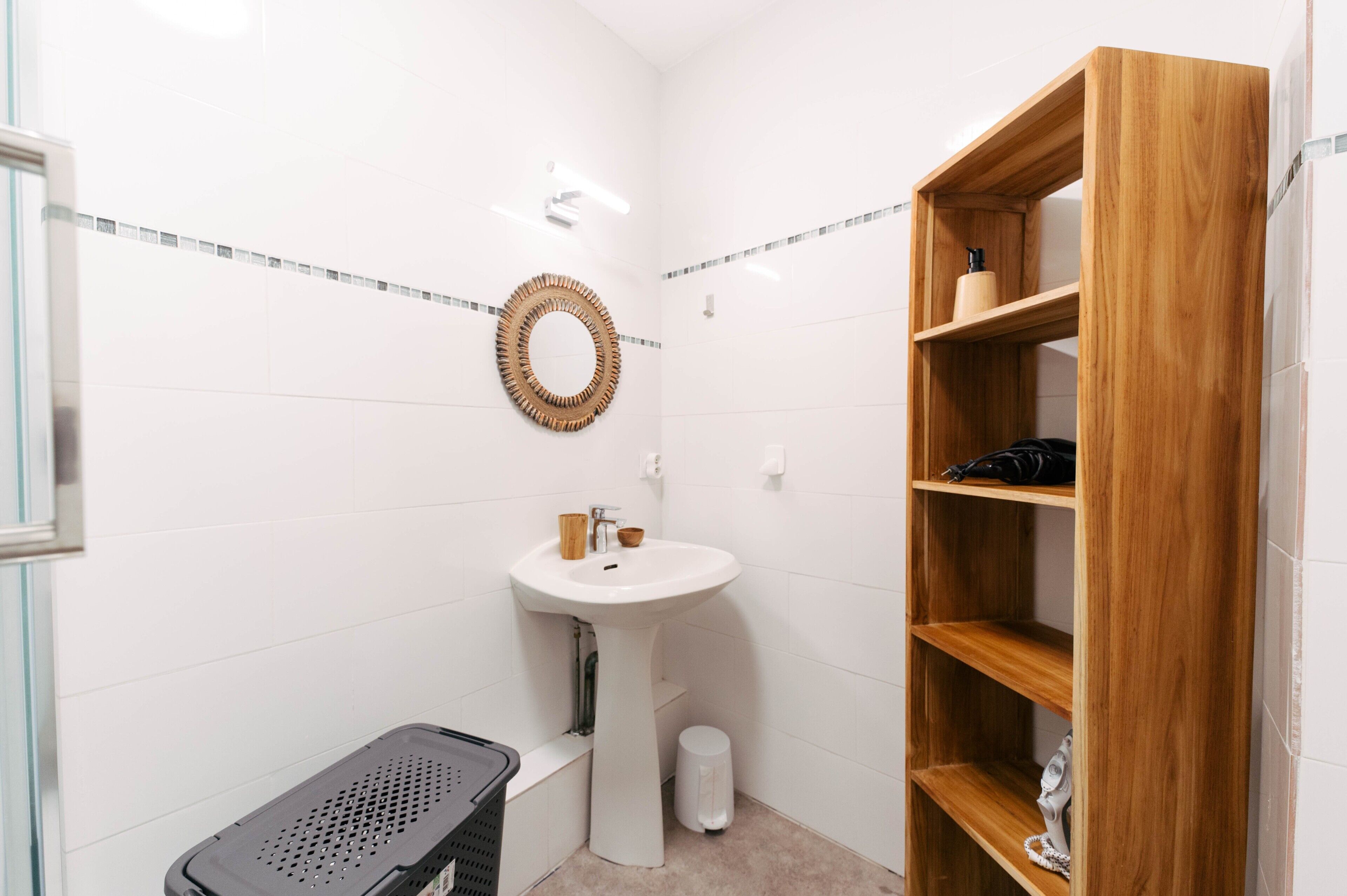 A bathroom with a pedestal sink, a wooden shelf, a trash bin, and a mirror.